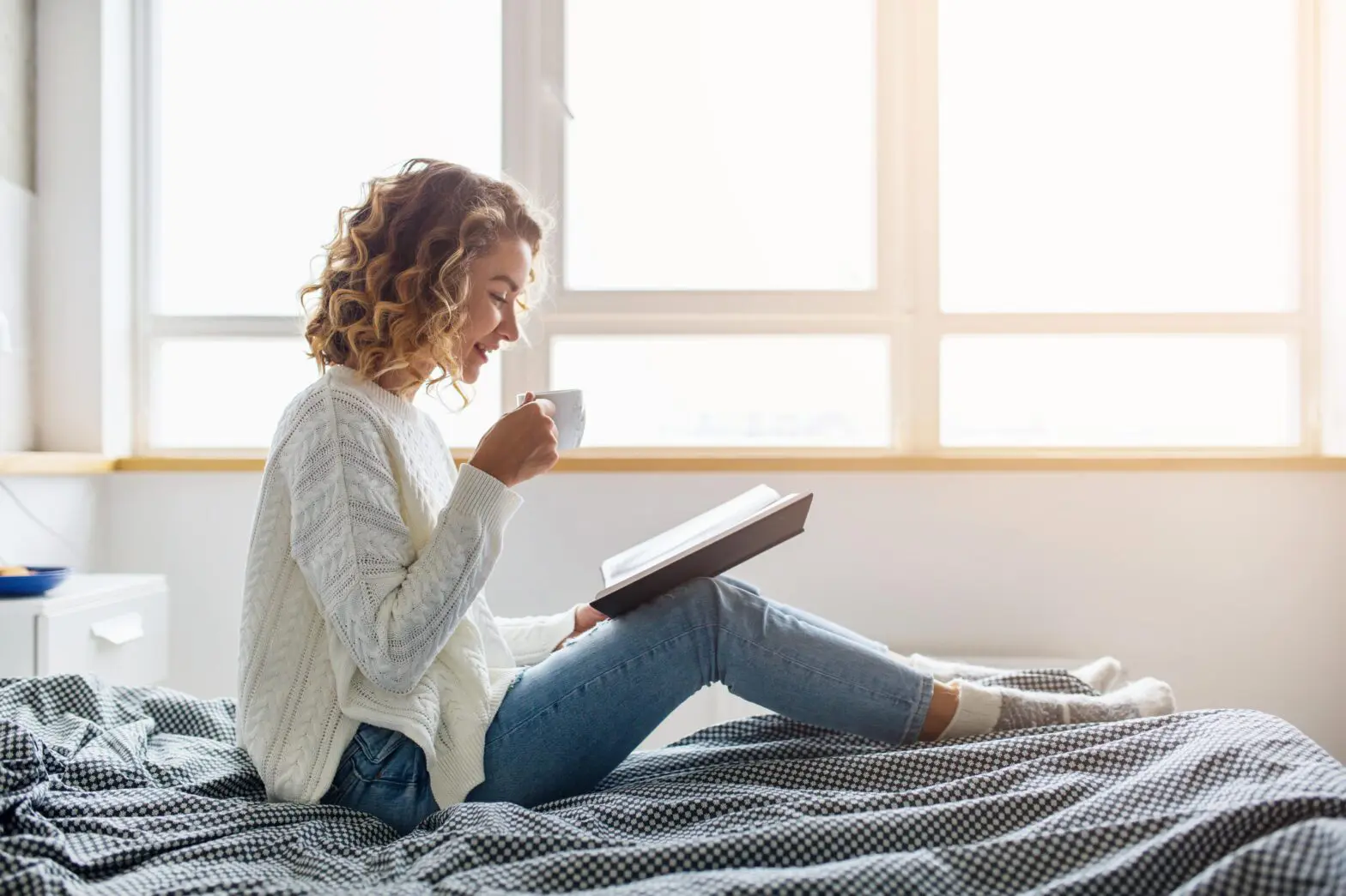attractive woman sitting on bed in the morning drinking tea reading book curly hair casual style blue jeans white sweater feeling comfortable at home having rest smiling Seven strategies to get good sleep during withdrawal