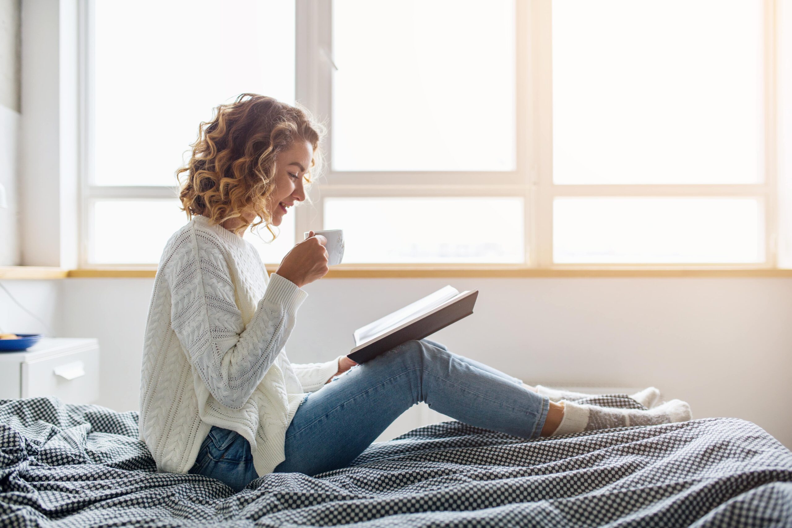 attractive woman sitting on bed in the morning drinking tea reading book curly hair casual style blue jeans white sweater feeling comfortable at home having rest smiling scaled Seven strategies to get good sleep during withdrawal